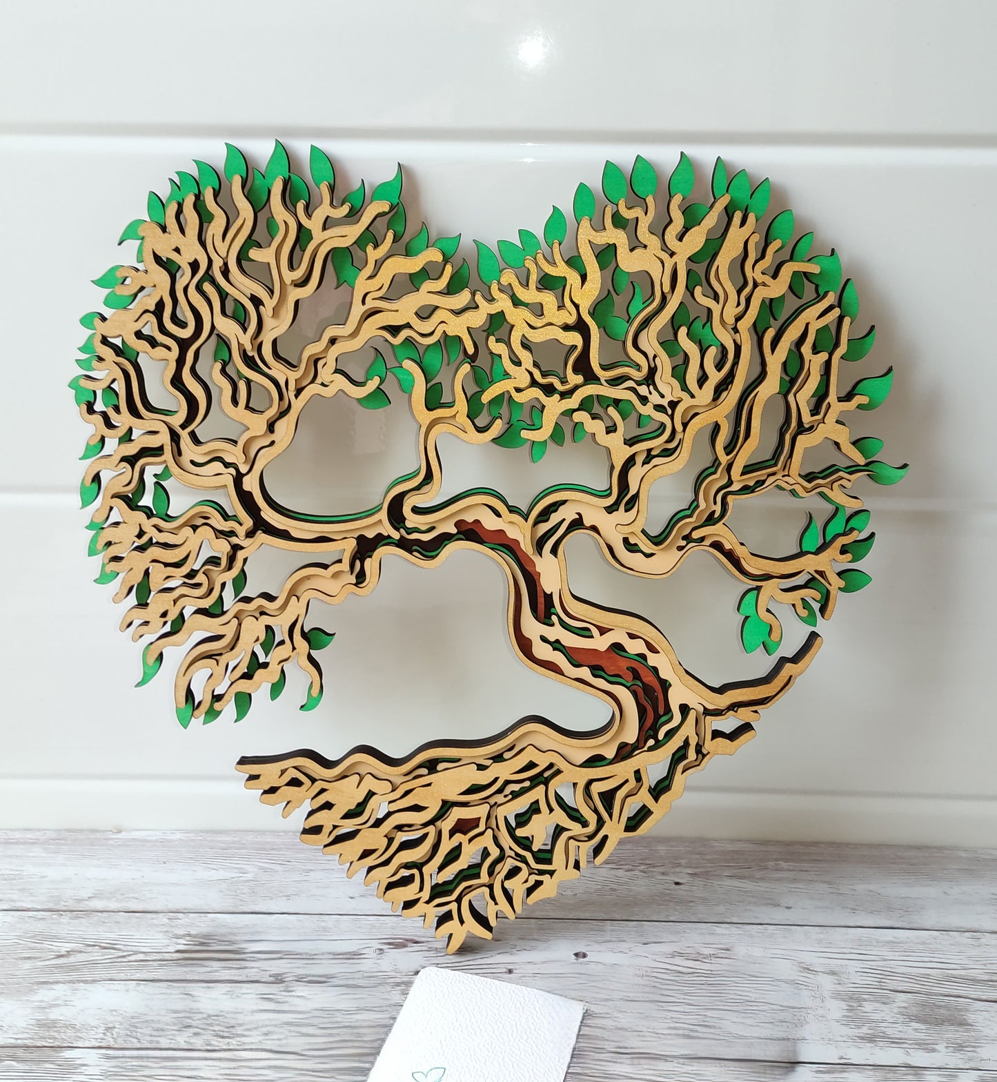 Décoration murale artisanale Arbre de Vie en forme de cœur, réalisée en 5 couches de bois de tilleul avec des détails de feuilles vertes.