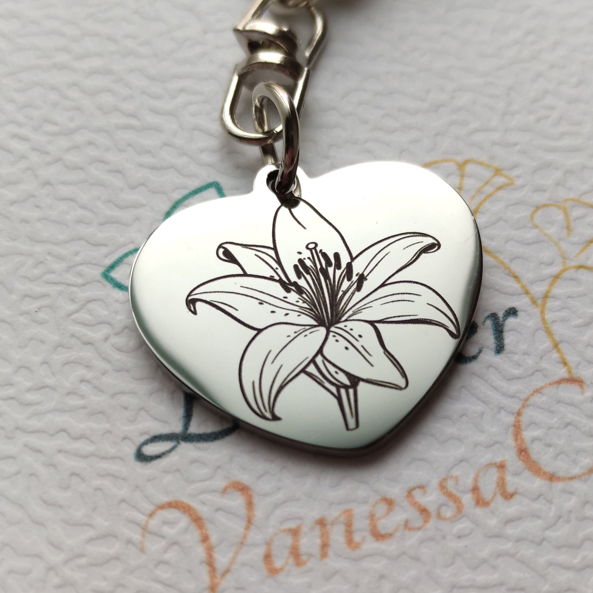 Porte-clés en forme de cœur en acier inoxydable poli miroir avec gravure laser d'une fleur de lys et texte personnalisé.