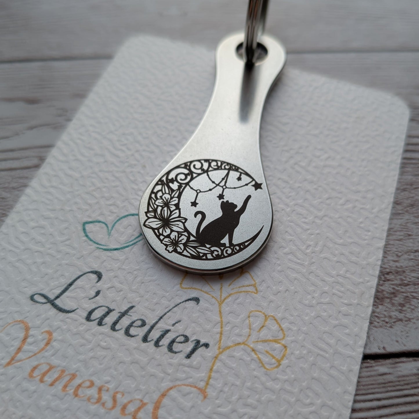 Porte-clés jeton de caddie amovible en acier inoxydable avec une gravure personnalisée représentant un chat sur un croissant de lune fleuri, posé sur sa carte de présentation L'Atelier VanessaCréa.