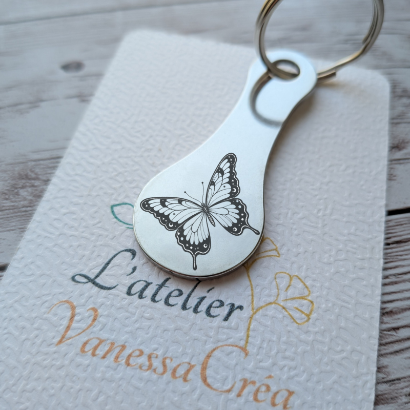 Porte-clés jeton de caddie en acier inoxydable gravé d'un motif papillon délicat, présenté sur son support L'Atelier VanessaCréa.