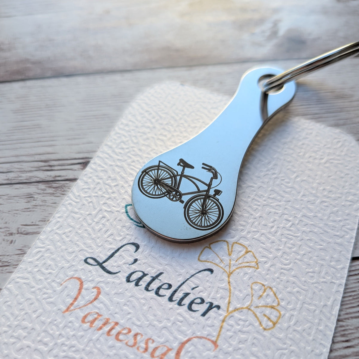 Porte-clés jeton de caddie en acier inoxydable gravé d'un motif de vélo rétro, posé sur le support de L'Atelier VanessaCréa.